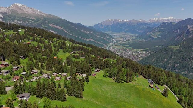 Idyllic small swiss village on hilltop of Alps. Aerial wide shot. Sunny day in summer. Snowy peak of alp mountains in distance. Green uphill landscape.