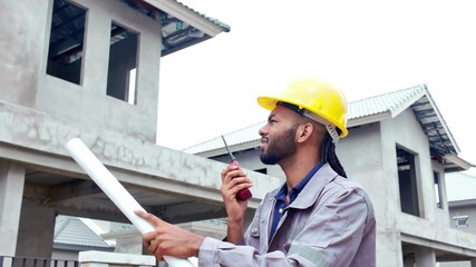 Male Construction worker or Engineer or Foreman stands at a building site holding blueprints and using a walkie-talkie radio to coordinate and consulting with the team.