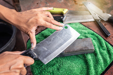 Sharpening knives with grindstone sharpener. Kitchen tools for use. Woman hand sharpens cleaver with whetstone on a green nano cloth pad on old table.