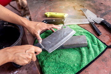 Sharpening knives with grindstone sharpener. Kitchen tools for use. Woman hand sharpens cleaver with whetstone on a green nano cloth pad on old table.