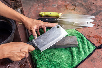 Sharpening knives with grindstone sharpener. Kitchen tools for use. Woman hand sharpens cleaver with whetstone on a green nano cloth pad on old table.