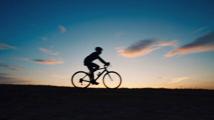 Obraz premium Silhouette Cyclist's Journey: A lone cyclist journeys onward, silhouetted against a breathtaking, multicolored sunset, capturing the spirit of exploration and the beauty of the open road.