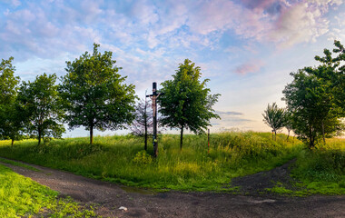 A solitary cross stands along a dirt road in the Czech countryside, symbolizing faith and tradition amidst open fields and rolling hills.