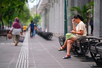A woman with a ponytail sits on a bench in a public area, smiling as she looks at her phone