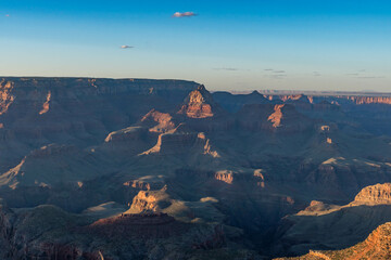 A breathtaking Grand Canyon sunset reveals layers of rugged red rock formations bathed in warm light and long shadows, while deep canyons and ridges form a mesmerizing view under a clear sky.