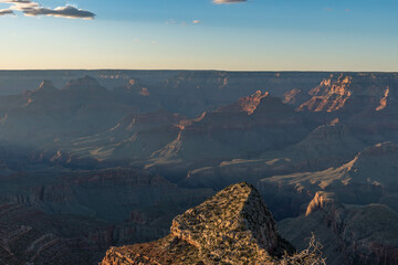 A breathtaking Grand Canyon sunset reveals layers of rugged red rock formations bathed in warm light and long shadows, while deep canyons and ridges form a mesmerizing view under a clear sky.