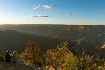 A breathtaking Grand Canyon sunset reveals layers of rugged red rock formations bathed in warm light and long shadows, while deep canyons and ridges form a mesmerizing view under a clear sky.