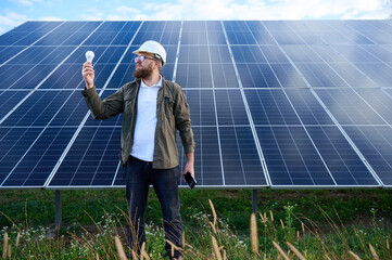 Engineer in uniform with protective helmet near solar station holding light bulb. Supervisor is...