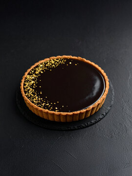 A glossy chocolate tart with a smooth ganache filling sits on a dark, textured surface. Its golden-brown crust contrasts beautifully with the deep chocolate tones, accented by delicate gold edible bea