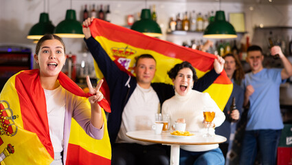 Group of sports supporters cheering for favorite team with flag of Spain while watching match game on TV in bar