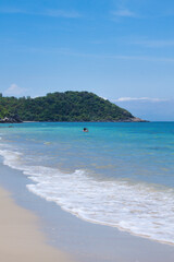 Tropical beach scene with turquoise waves gently washing ashore, a lone swimmer in the sea, and distant islands under a clear blue sky. 
