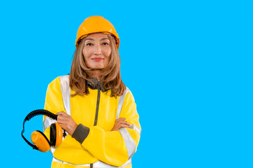 Construction worker stands confidently wearing a safety helmet and headphones against a bright blue background in a construction setting