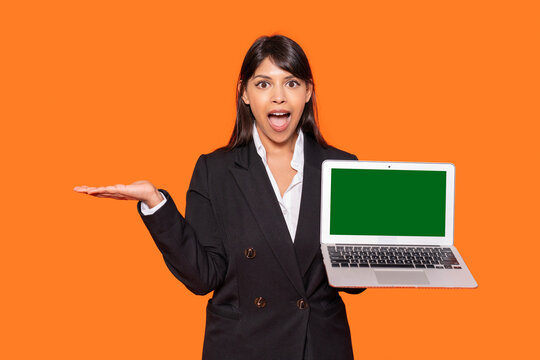 Young woman in a black suit excitedly holds a laptop with a green screen against an orange background while gesturing with her free hand - Powered by Adobe