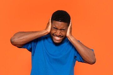 Person expressing discomfort while covering ears against loud noise in a bright orange background,...