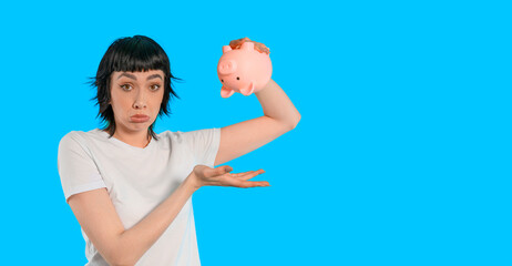 Young woman holds piggy bank looking frustrated about finances in a bright blue background during a casual indoor moment