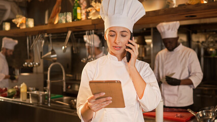 Portrait of busy professional female caucasian chef using tablet online and talking on phone to...