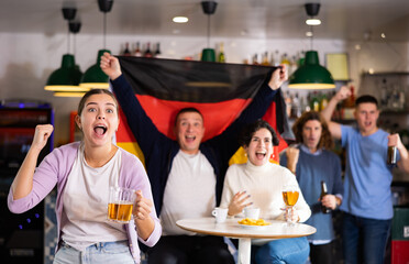 Company of emotional young adult fans supporting German team with state flag while drinking beer in sports bar