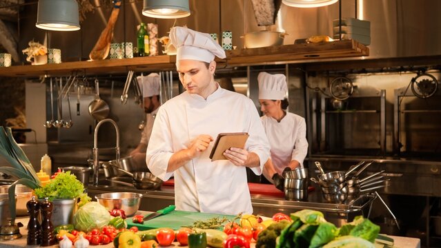 Portrait of attractive professional chef looking at tablet searching for recipe for salad. Handsome young cook cooking at modern restaurant kitchen with fresh vegetables and ingredients.