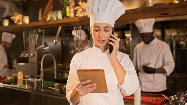 Portrait of busy professional female caucasian chef using tablet online and talking on phone to order ingredients delivery. Attractive cute successful woman cook wearing white uniform.
