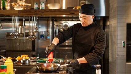Portrait of beautiful professional male asian chef in black uniform and gloves frying and cooking delicious dishes using pan. Attractive skillful cook mixing vegetables and adding sauce.