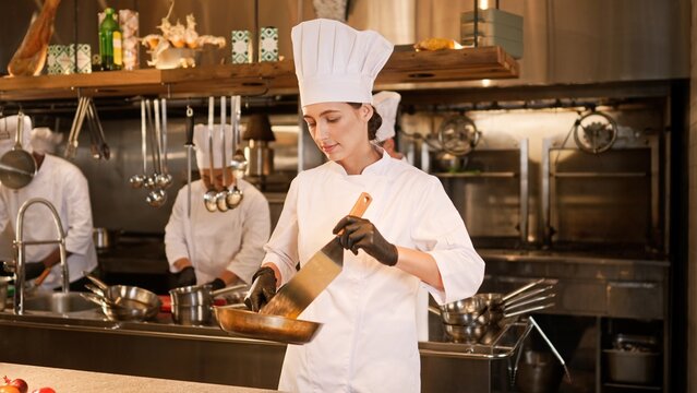 Portrait of focused beautiful female caucasian chef frying delicious food on pan and mixing. Attractive skillful cook in white uniform cooking tasty dishes at modern kitchen of fancy restaurant.
