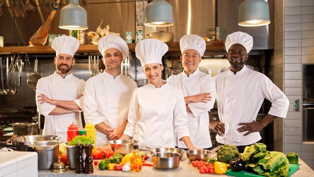 Portrait of professional successful multi-ethnic team of cooks standing in modern kitchen posing looking at camera smiling. Happy caucasian, african american and asian male and female chefs.