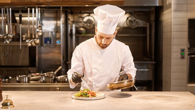 Portrait of skillful professional caucasian chef in white uniform and gloves decorating delicious salad adding shrimps. Handsome cheerful male cook with beard enjoying cooking at kitchen. - Powered by Adobe