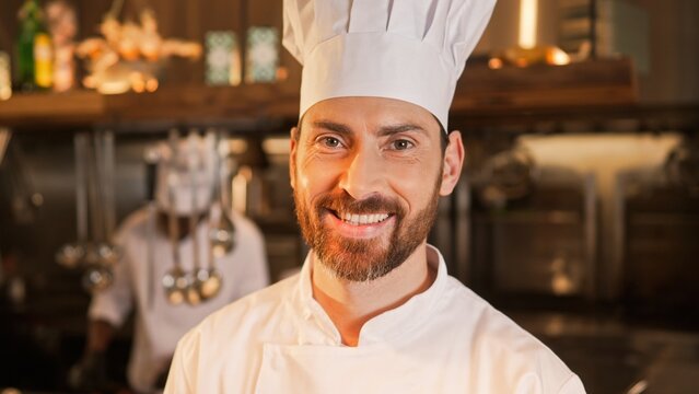 Close-up portrait of famous handsome young caucasian chef looking at camera smiling. Beautiful male cook wearing uniform working in kitchen of fancy restaurant. Cooking concept.