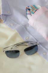 Stylish featuring a pastel-colored men’s shirt with a folded pocket square in soft tones. Transparent-frame sunglasses with dark lenses rest on the fabric, creating a refined and summery fashion.