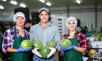 Smiling factory workers holding ripe whole watermelons and looking at camera during sorting ripe...