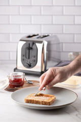 Slice of toast being buttered by hand, with butter dish, strawberry jam, and metallic toaster in the background. Bright kitchen countertop scene. Copy space. Concept of homemade breakfast 