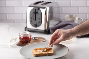 Male hand spreading butter on a slice of toast on a ceramic plate. Strawberry jam, toaster, and a grey napkin complete the morning kitchen scene. Copy space. Concept of  natural breakfast at home