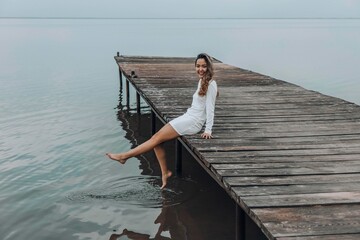 Asian woman in a white dress, sitting on the edge of wooden pier, dipping her feet into calm water...