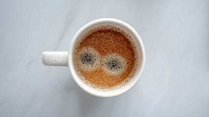 A top-down view of a cup of coffee with a creamy froth pattern that resembles a pair of wide, cartoon-like eyes. The white ceramic mug sits on a light, smooth surface.