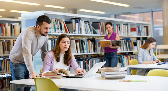 Two friendly young adult male and female students preparing for exam together in university library, using textbooks and laptop