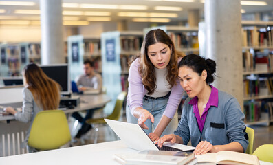 Two positive female colleagues working together on joint project in library, looking for information in books and Internet..
