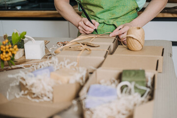 Person writes on ribbon-wrapped kraft boxes near handmade soaps and shredded filler. Handmade soap branding, natural skincare sets, self-care packaging, organic beauty business