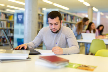 Focused guy student studying at the university studies in the library on a laptop while preparing...