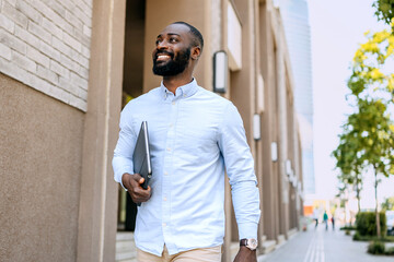 Young black man walking down a city street with a laptop under his arm during a sunny day