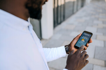 Man checking weather forecast on smartphone while standing outdoors in urban setting during sunny...