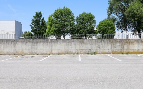 Surrounding concrete wall with metal grid railing on top. Industrial building, trees and sky on behind, empty parking lot in front. Background for copy space.