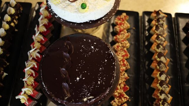 Top view of glossy chocolate cake and coconut dessert surrounded by rows of mini layered pastries and fruit bars creates a luxurious and festive dessert table perfect for hotel events and receptions