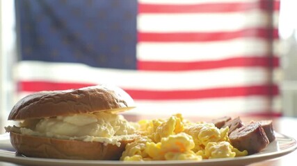 A plate with a bagel sandwich and scrambled eggs in front of an American flag. The scene captures a classic American breakfast setting.