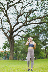 Young sportswoman wiping sweat with towel after exercising in a green park with big trees, listening music with headphones, enjoying a healthy lifestyle