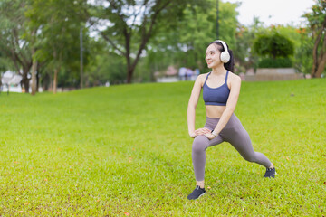 Young Asian sportswoman wearing headphones, stretching legs in a park, enjoying her workout with music, promoting healthy lifestyle and wellness