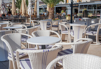 A sunny outdoor cafe terrace with white wicker furniture, striped cushions, and closed umbrellas, set in a Mediterranean plaza with palm trees and flower planters.