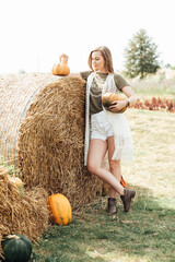 young woman stands at pumpkin festival, holding large orange and green pumpkin. She is surrounded by rows of pumpkins and a wooden display. The autumn farm setting captures the harvest season vibe.