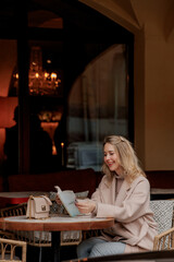 A young Caucasian woman with blonde hair sits at a cafe table in Saint Petersburg, reading a book. The background features elegant decor and warm lighting.