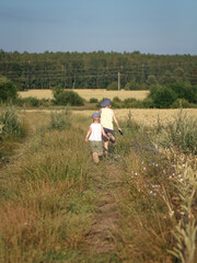 Two young children walking along a narrow path through a rural summer field. The kids are seen from behind, wearing casual summer clothes and hats, surrounded by tall grass, wildflowers.