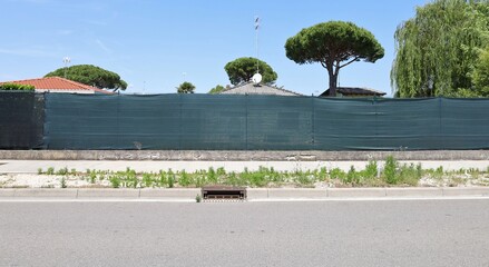 Green tarpaulin on vinyl coated fence at the roadside. House roofs, trees and sky on behind,...